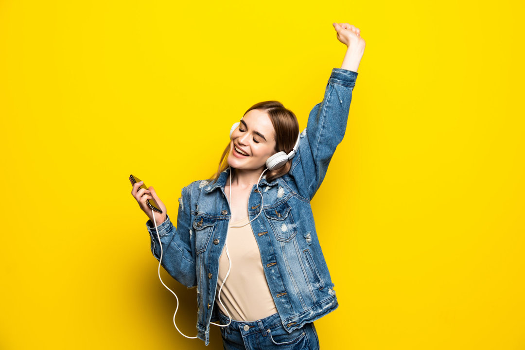 Happy cheerful woman wearing wireless headphones listening to music from smartphone studio shot isolated on yellow background