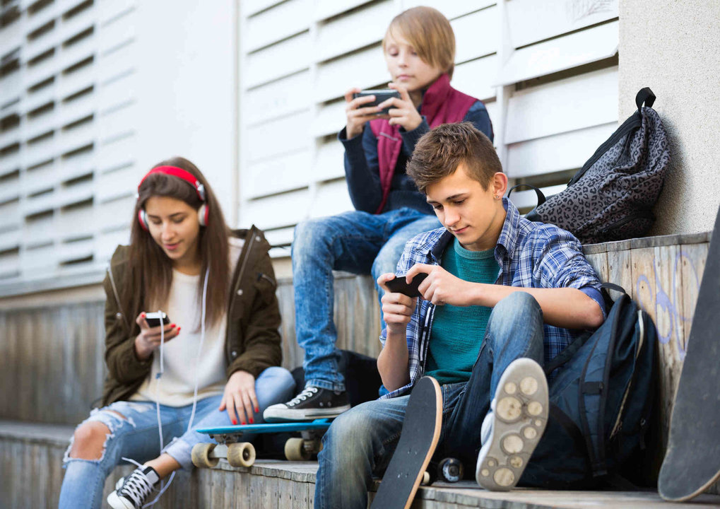Two teenage males and their girlfriend relaxing with mobile phones outdoor. Focus on guy