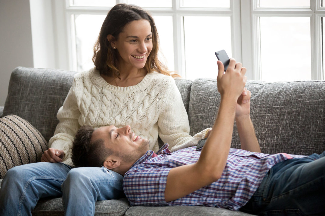 Smiling happy millennial couple using smartphone relaxing on couch, boyfriend and girlfriend checking social network news online on cell together at home, young man showing woman new mobile phone app